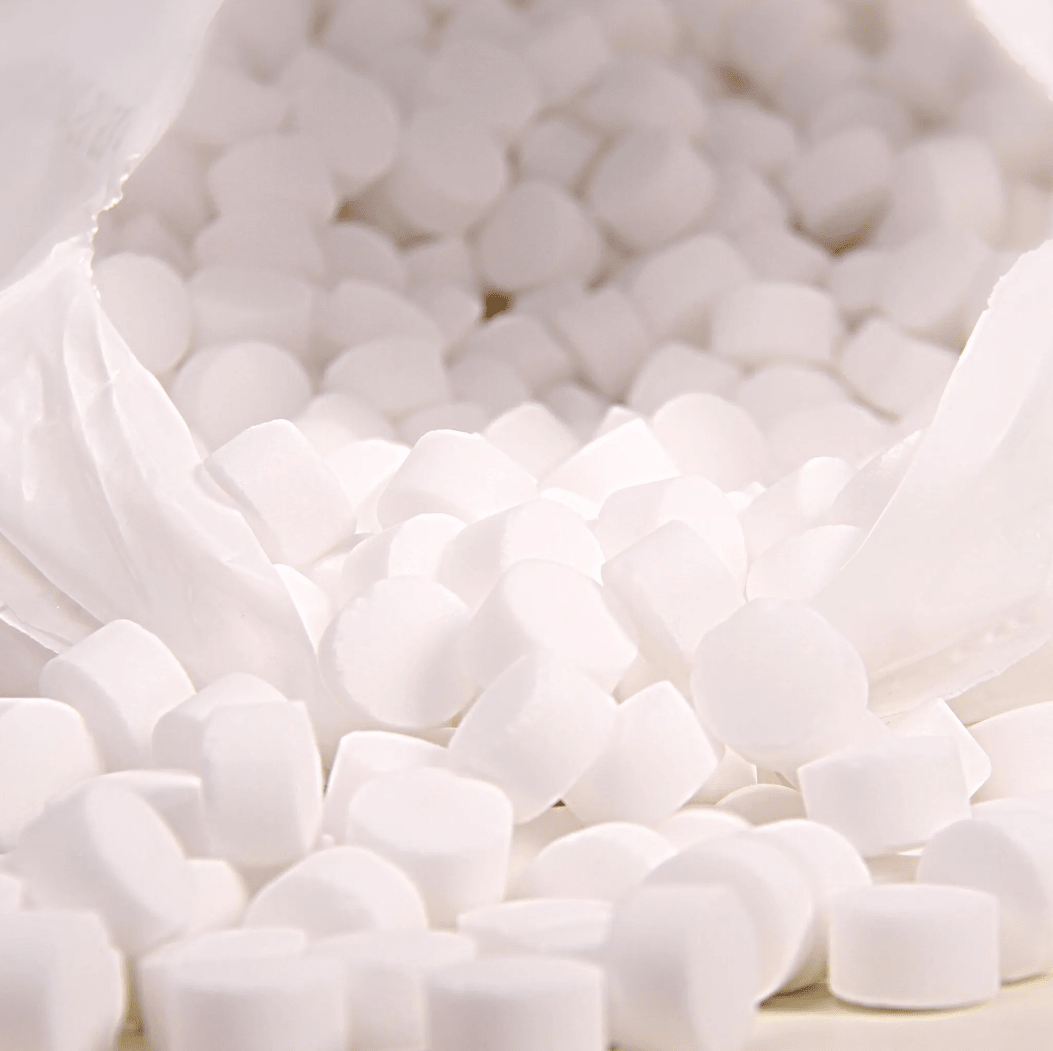 En närbild visar en vit plastpåse med texten ”K+S Minerals and Agriculture GmbH”, som innehåller Regenit-koktsaltstabletter enligt EN 973 typ | säck (25 kg), vilka faller ner på en yta. De små, runda tabletterna har en enhetlig form och storlek och är perfekta för användning i vattenavhärdningsanläggningar.