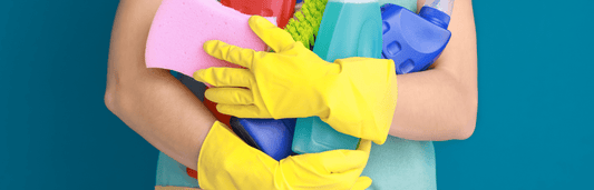 Person mit gelben Reinigungshandschuhen hält Schwamm, Bürste und Reinigungsflaschen vor blauem Hintergrund in Nahaufnahme