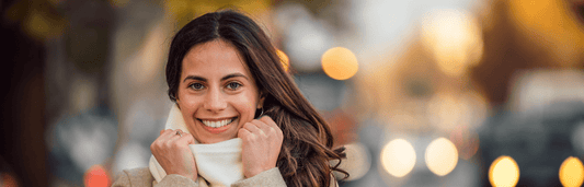 Lächelnde Frau mit Schal hält diesen am Hals fest, unscharfer Hintergrund mit Lichtern in warmer Abendstimmung