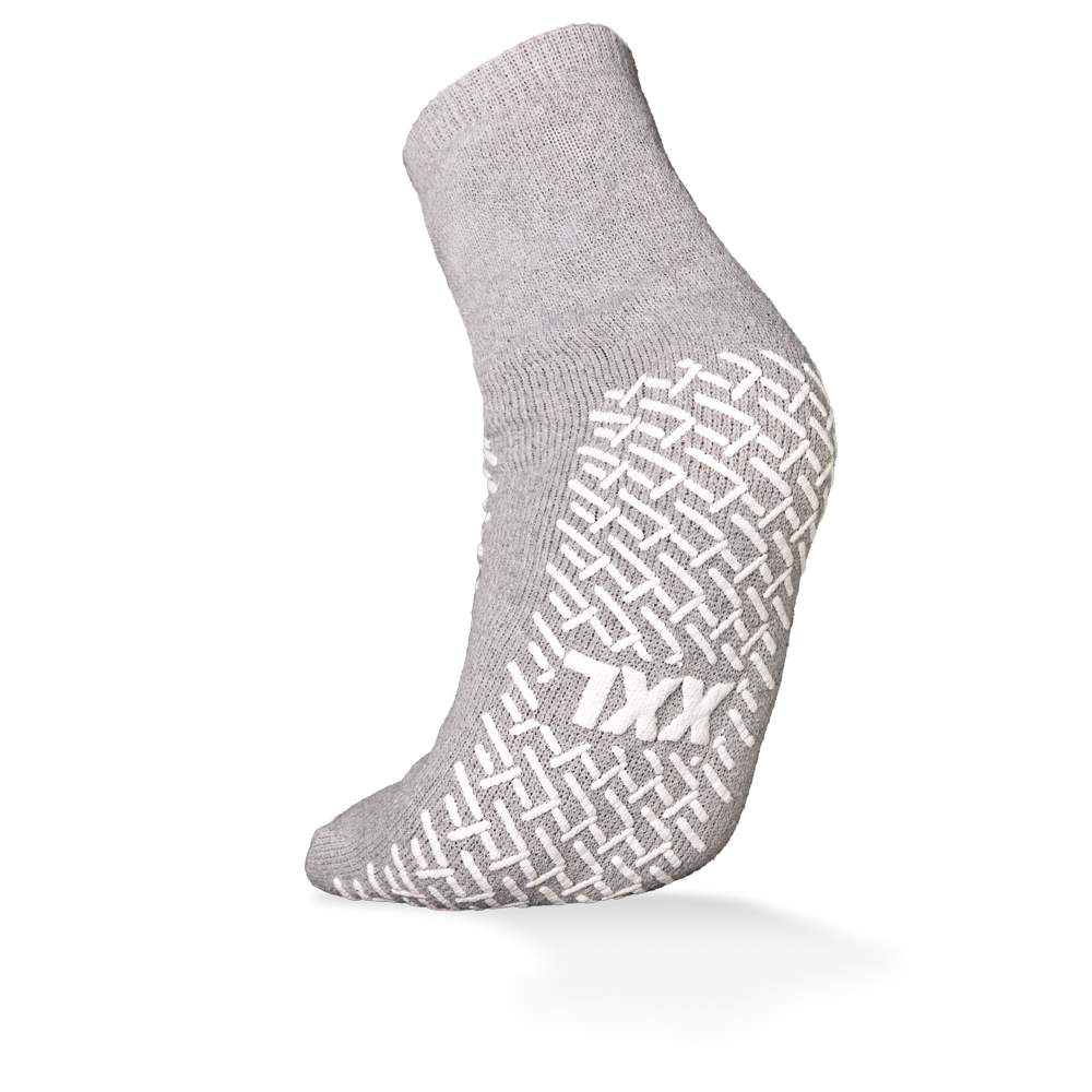 Abgebildet ist eine einzelne graue ECO-PLUS Anti-Rutsch Einwegsocke der AMPri Handelsgesellschaft mbH mit weißem Anti-Rutsch-Griff und "TXX"-Schriftzug auf der Sohle. Beidseitig beschichtet, ideal für medizinische Bereiche; der Hintergrund ist schlicht weiß.