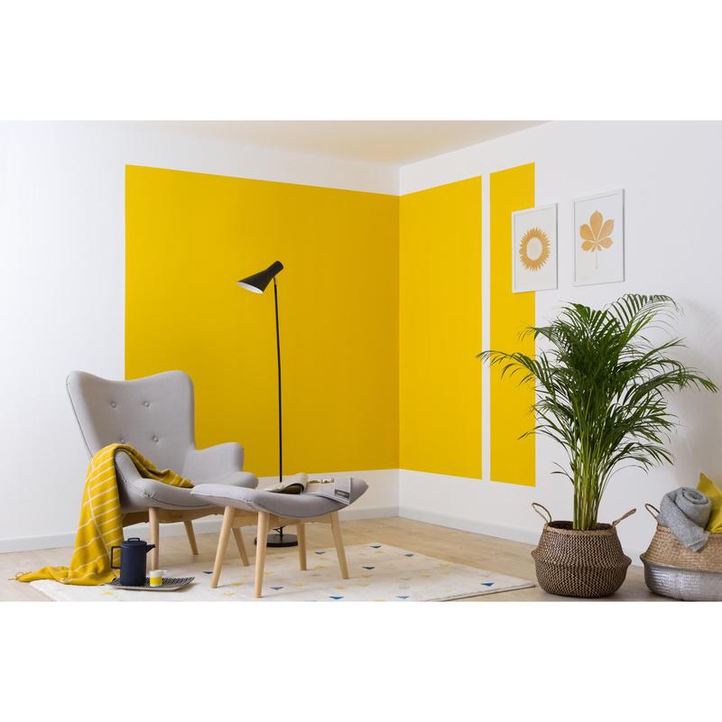 Ett modernt vardagsrum med en grå fåtölj, en ottoman, en svart golvlampa, en gul filt och en krukväxt. De kraftiga gula geometriska formerna på väggen skapades med 3M Deutschland GmbH ScotchBlue™ målartejp för skarpa linjer.