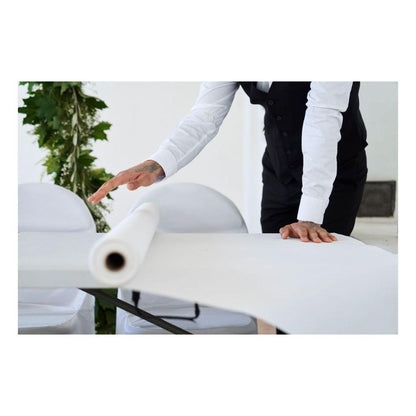 Eine Person in einem weißen Hemd und einer schwarzen Weste rollt eine Dunicel-Tischdeckenrolle der Duni GmbH (1,18 m x 10 m) über einen Tisch aus, im Hintergrund sind weiße Stühle und grünes Laub zu sehen.