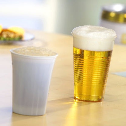 Två ölglas står på ett ljust träbord - ett i en klar Starpak 75 dricksmugg, PS klar med skumkant från Starpak, som visar gyllene öl, det andra i en ogenomskinlig vit mugg. I bakgrunden syns suddig mat.
