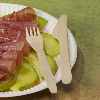 En pappersplatta med skivade gröna äpplen och rullat saltat kött vilar tillsammans med en PAPSTAR träkniv "pure" 16,5 cm från PAPSTAR GmbH och en trägaffel på en grön yta.