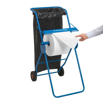 En blå pappershanddukshållare med en stor vit Scott® ESSENTIAL™ läkarroll (6 x 200 ark) från Kimberly-Clark GmbH understryker hygienen; en svart sopsäck är i närheten, medan en persons hand drar ett ark från rullen.