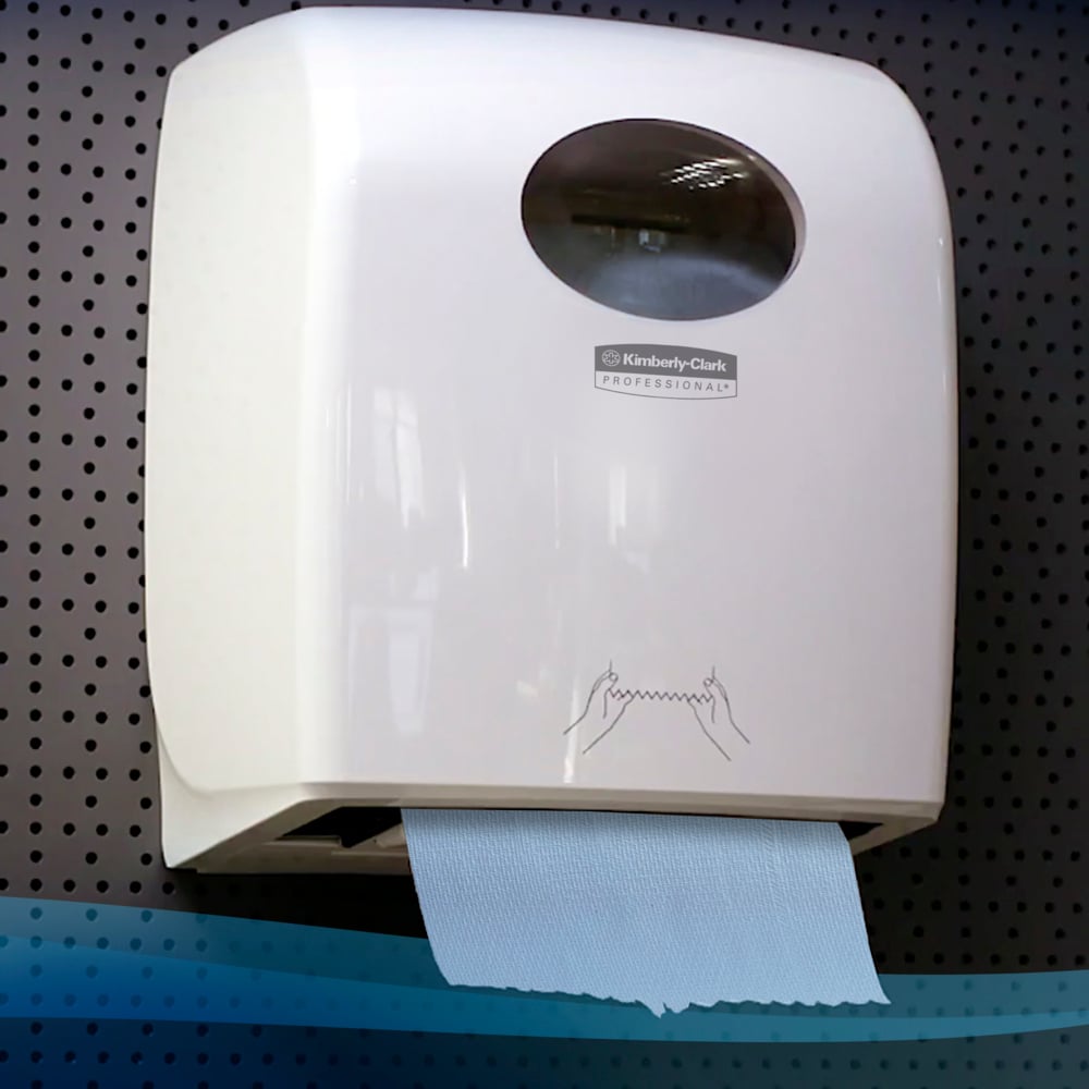 Ein weißer, an der Wand montierter Spender der Kimberly-Clark GmbH hält eine Scott® Handtücher - Rolle (304,00 m x 19,80 cm) mit einem blauen Handtuch und einer einfachen, illustrierten Anleitung vor einem dunklen Hintergrund aus Lochblech.