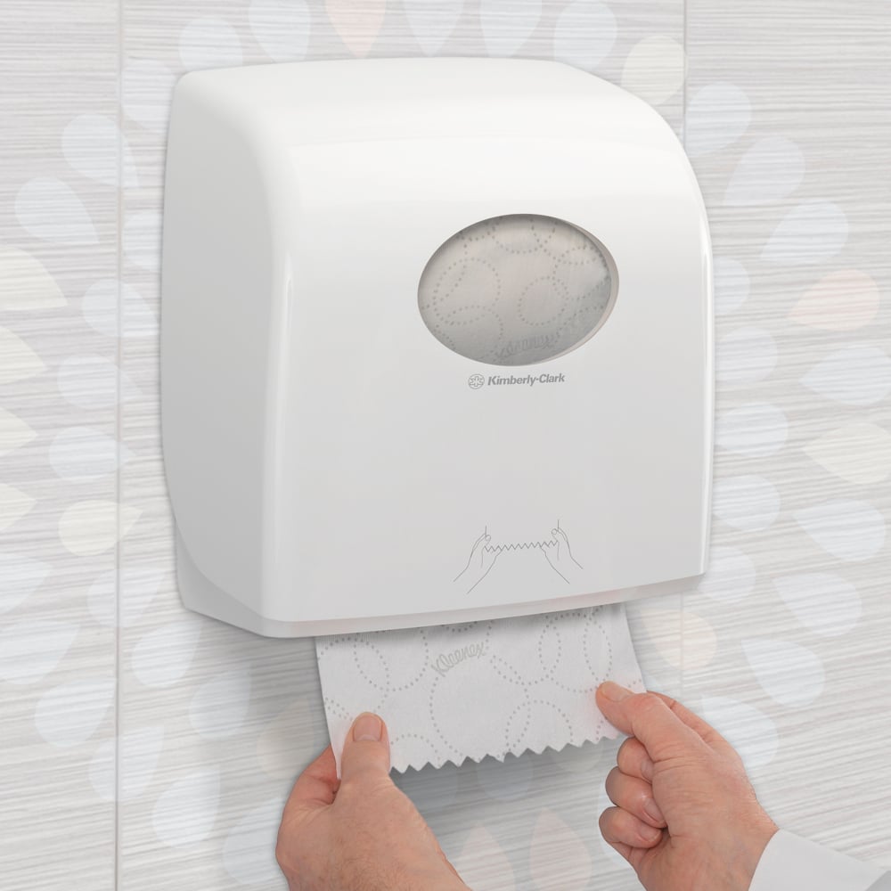 Någon drar ut en Kleenex® Ultra™ Slimroll™ pappershandduk från en vit, väggmonterad dispenser med fönster och symboler i en kaklad toalett. Produkt: 6-rullars kartong från Kimberly-Clark GmbH.