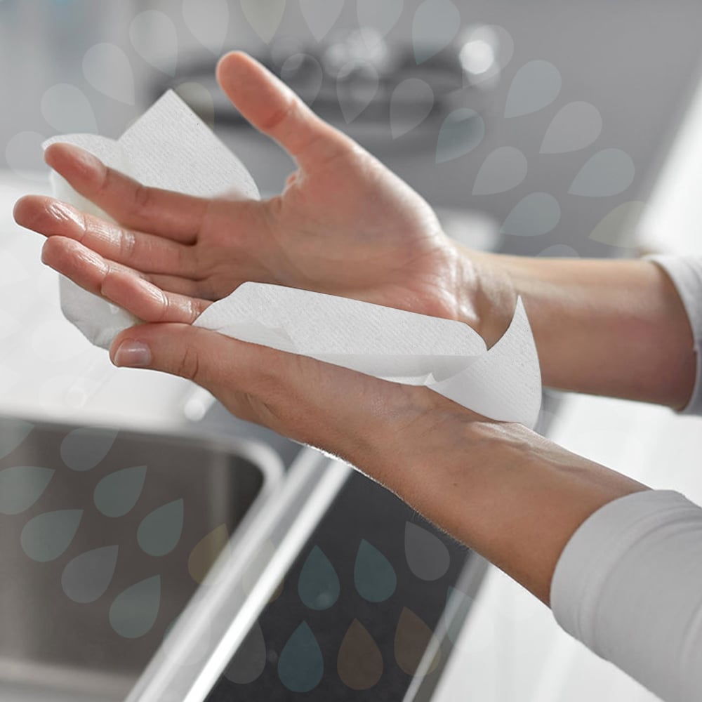 En person torkar sina händer över ett handfat med Kleenex® vikta handdukar - Vit/Storförpackning (15 clips x 124 ark | kartong) från Kimberly-Clark GmbH, där båda händerna och en del av handfatet syns i bakgrunden.