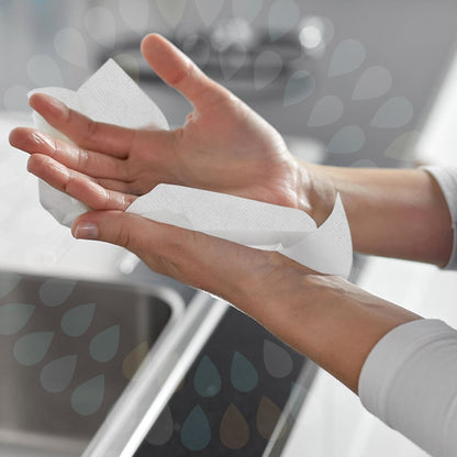 En person torkar sina händer över ett handfat med Kleenex® vikta handdukar - Vit/Storförpackning (15 clips x 124 ark | kartong) från Kimberly-Clark GmbH, där båda händerna och en del av handfatet syns i bakgrunden.