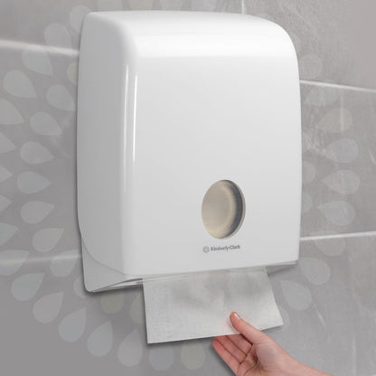En hand drar ut en Kleenex® Ultra™ handduk från en väggmonterad vit dispenser från Kimberly-Clark GmbH, som är installerad på en kaklad vägg (Interfold/Vit/Stor - 30 klämmor x 94 ark | Kartong, 30 paket).