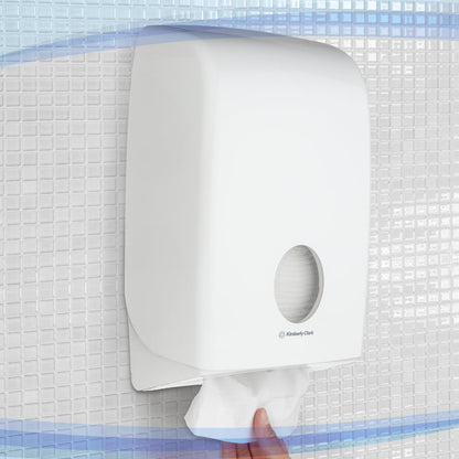 En hand drar ut en pappershandduk från den vita Aquarius™-dispensern för vikta handdukar från Kimberly-Clark GmbH, som är monterad på en kakelvägg. Dispensern har ett fönster för hygienisk, enkel handduksutmatning.