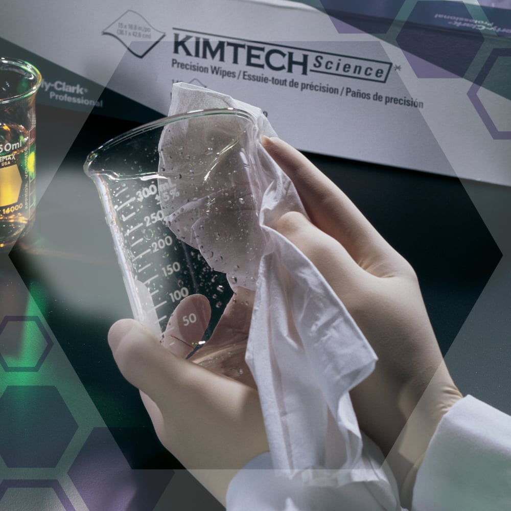 En person med vita handskar torkar av ett klart glasbägare med Kimtech® Science precisionsservetter från Kimberly-Clark GmbH. I bakgrunden står en låda med servetterna (100 ark, 2-lagers) och en bägare med vätska.