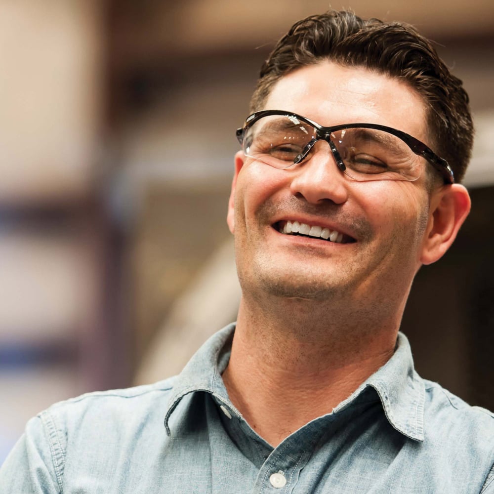Ein Mann in einem hellblauen Button-up-Hemd lächelt, während er Auslaufartikel der Kimberly-Clark GmbH trägt: KleenGuard® V30 Nemesis™ Schutzbrille mit Beschlagfrei-Beschichtung; der sanft verschwommene Hintergrund sorgt für Aufmerksamkeit.
