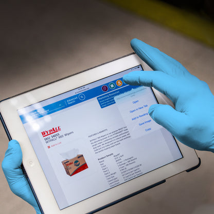 Eine Person, die KleenGuard® G10 Comfort Plus Blaue Nitril-Handschuhe von Kimberly-Clark GmbH trägt, hält ein Tablet, das eine Produktseite mit Bild, Beschreibung und Kontextmenü zeigt; der Hintergrund ist unscharf.