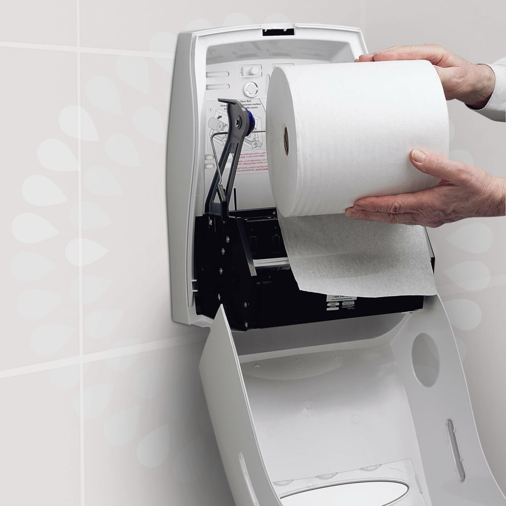 En person laddar en Kleenex® Ultra™ handduksrulle från Kimberly-Clark GmbH i en vit, öppen väggdispenser på en toalett för att säkerställa hygienisk handtorkning. På dispenserns lock finns en instruktion tryckt.