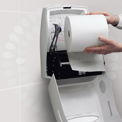 En person laddar en Kleenex® Ultra™ handduksrulle från Kimberly-Clark GmbH i en vit, öppen väggdispenser på en toalett för att säkerställa hygienisk handtorkning. På dispenserns lock finns en instruktion tryckt.