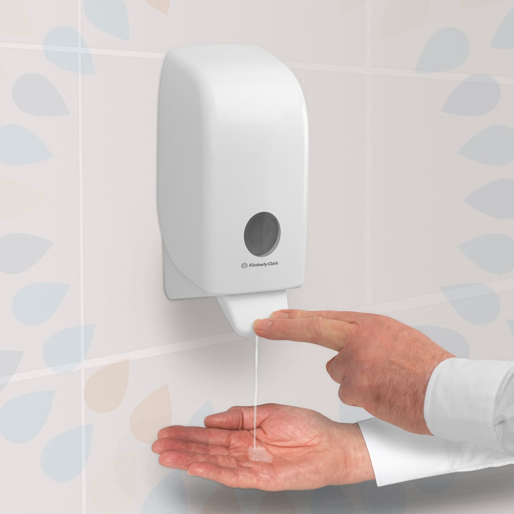 En person trycker på en transparent Kleenex® Extra Gel Handdesinfektionsdispenser från Kimberly-Clark GmbH, som är monterad på en kaklad vägg, och dispenserar handdesinfektionsgel från en 1-liters kassett på deras öppna handflata.