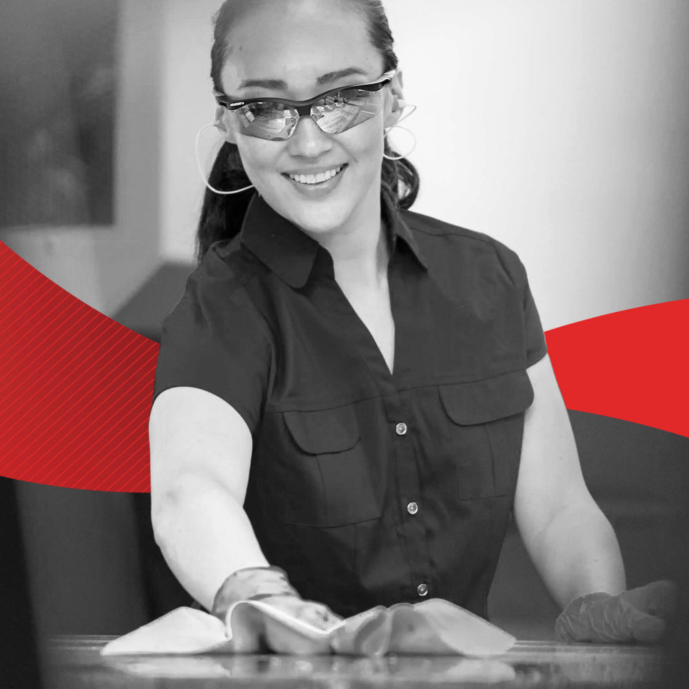 Eine Frau mit Handschuhen und Brille lächelt, während sie eine Oberfläche mit einem WypAll® X60 General Clean - BRAG™ Box Tuch (41,80 × 28,30 cm) der Kimberly-Clark GmbH reinigt; im Hintergrund erscheint ein rotes Grafikdesign.