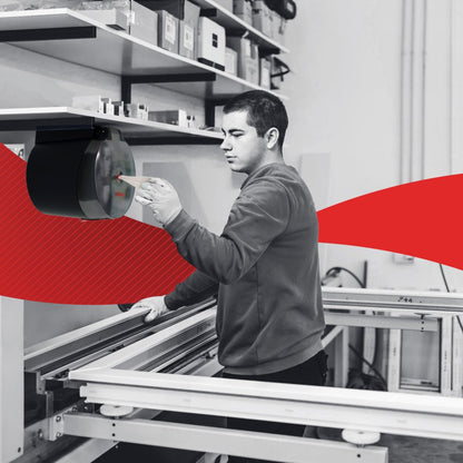 En person använder en väggmonterad WypAll® Reach Plus™ Reach™ Centrefeed-dispenser från Kimberly-Clark GmbH på en arbetsplats. Bakom finns hyllor med kartonger och andra föremål, medan en röd grafik korsar bilden horisontellt.