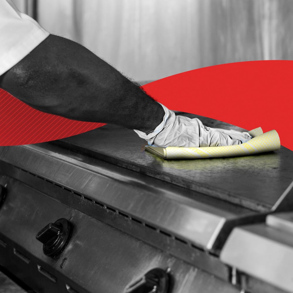 En person torkar av en kommersiell köksspis med handskar med en gul WypAll® X80 Critical Clean - Interfold (41 x 33 cm) från Kimberly-Clark GmbH; i bakgrunden visas ett rött grafiskt element.