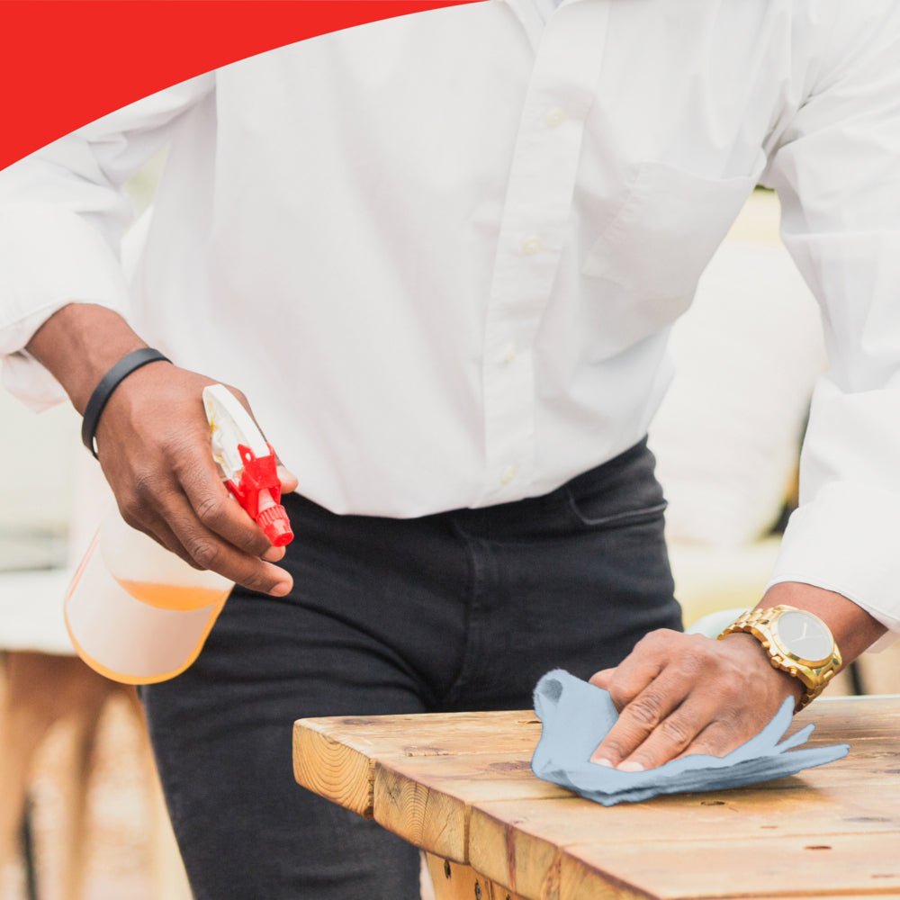 En person med vit skjorta och klocka rengör ett träbord med en blå duk, möjligen WypAll® L20 General Clean - BRAG™ Box från Kimberly-Clark GmbH. Bilden har en röd böjd kant upptill som betonar uppmärksamheten på detaljer.