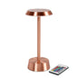 Den trådlösa NOUR LED-lampan i roséguld från Duni GmbH har en rund bas och platt ovansida för modern stil. Bredvid lampan ligger en fjärrkontroll med färgglada knappar.