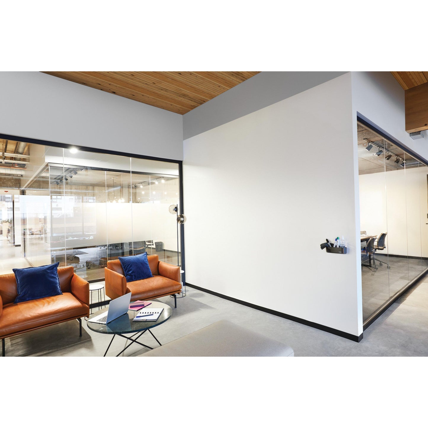 Ein moderner Büro-Wartebereich mit zwei braunen Ledersesseln, blauen Kissen, einem gläsernen Couchtisch mit Laptop und Notebook und gläsernen Besprechungsräumen, die mit der Post-it® Easy Erase Whiteboard-Oberfläche der 3M Deutschland GmbH ausgestattet sind.