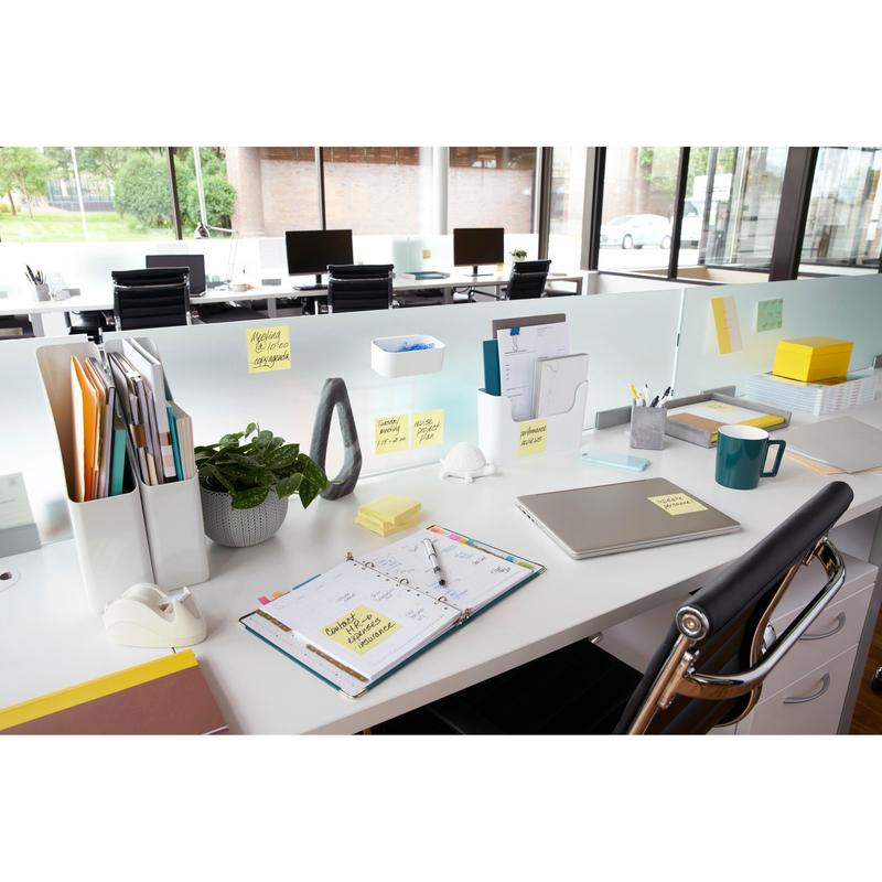 Ett modernt skrivbord med laptop, planerare, kaffekopp, växt, skrivmaterial och 3M Post-it® Notes Gult (76x76 mm, 100 blad/block), idealiskt för snabba anteckningar. Akterna är organiserade och stora fönster ger utsikt utåt; skrivborden är arrangerade i rad.