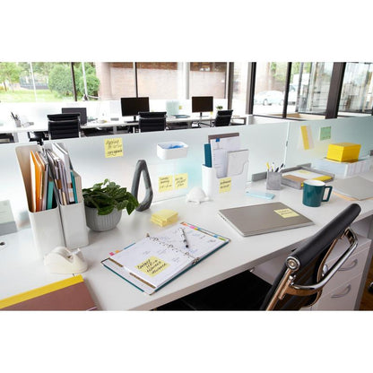 Ett modernt skrivbord med laptop, planerare, kaffekopp, växt, skrivmaterial och 3M Post-it® Notes Gult (76x76 mm, 100 blad/block), idealiskt för snabba anteckningar. Akterna är organiserade och stora fönster ger utsikt utåt; skrivborden är arrangerade i rad.
