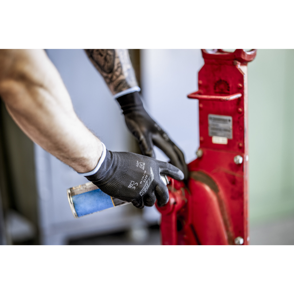 En person med NITRAS nylonskyddshandskar, svart, PU-belagda från AS Arbeitsschutz GmbH rengör eller smörjer med en blå sprayburk en röd maskindel av metall. Endast armar och händer syns.