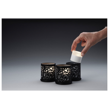 En hand placerar ett litet cylindriskt ljus i en av tre ljushållare Twine 75 x Ø 75 mm Svart från Duni GmbH, som är utrustade med detaljerade utskärningar och är idealiska för modern design. Bordsdekorationen är placerad mot en bakgrund med en färgövergång från mörk- till ljusgrå.