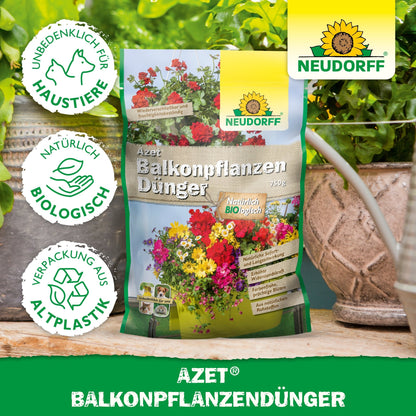 En påse Azet Balkongväxtgödsel från Neudorff Shop står på ett bord mellan krukväxter och balkonglådor. Förpackningen visar blommor, text och symboler för djurskydd, biologiska ingredienser och förpackningar av bioplast.