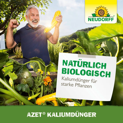 En gråhårig man står i en trädgård, ler och håller upp en gul zucchini med knuten näve. På en skylt står det "Naturligt biologiskt, Azet Kaliumgödsel för starka växter." Neudorff Shops varumärke är synligt.
