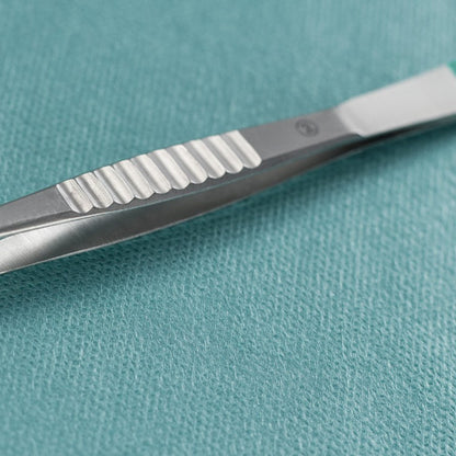 En närbild av en Paul Hartmann AG Hartmann Splitterpincett rakt 9cm, steril (förpackning med 40 stycken), som ligger bredvid en kirurgisk metallskalpell på en teal-färgad, strukturerad medicinsk duk.