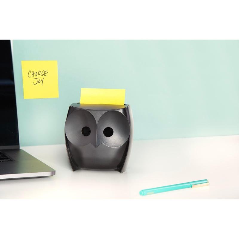 En svart Post-it® Z-Notes-dispenser "uggla" (3M Deutschland GmbH) med gula Super Sticky Z-Notes står på ett vitt skrivbord, bredvid en laptop och en blå penna. På väggen hänger en "CHOOSE JOY"-lapp mot en ljusblå bakgrund.