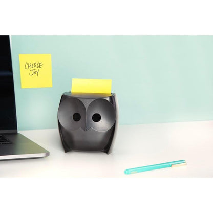 En svart Post-it® Z-Notes-dispenser "uggla" (3M Deutschland GmbH) med gula Super Sticky Z-Notes står på ett vitt skrivbord, bredvid en laptop och en blå penna. På väggen hänger en "CHOOSE JOY"-lapp mot en ljusblå bakgrund.