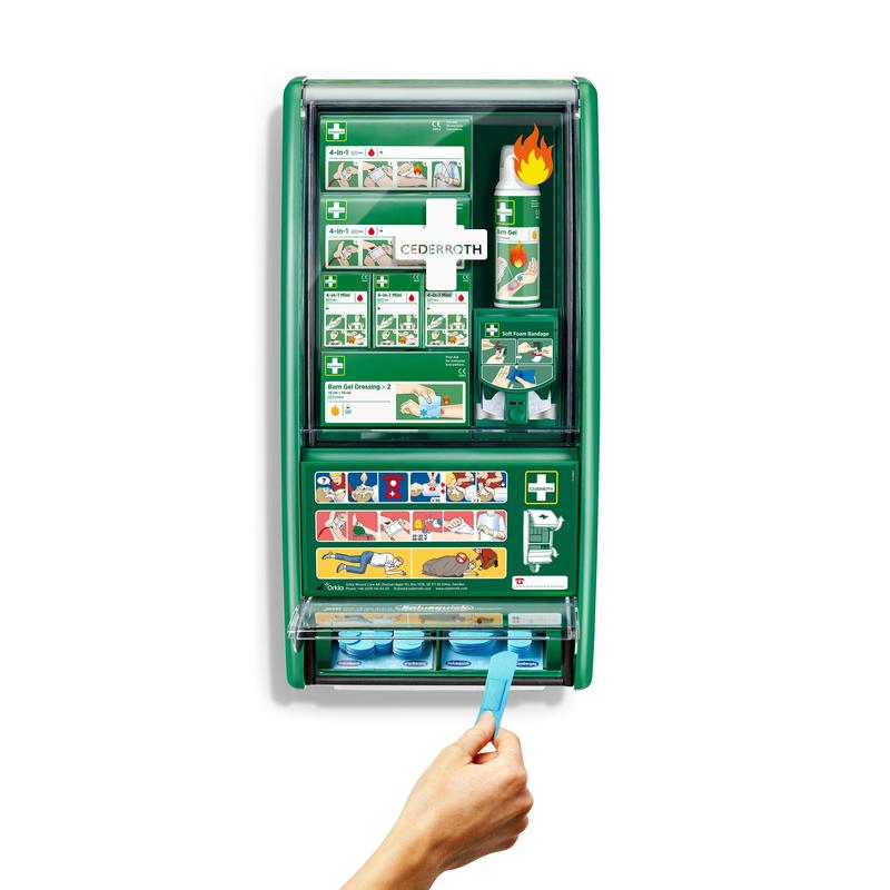 En grön Cederroth första hjälpen & brännskadestation från Orkla Wound Care AB är monterad på en vit vägg, utrustad med förband, brännskadegel, ögonskölj och mer. En hand drar ett blått klisterband ur den nedre dispensern av 1-kartongen.