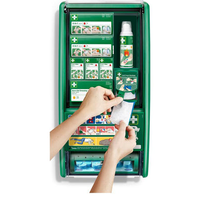 En person håller ett brännskadegel-förband framför Cederroth första hjälpen & brännskadestation (1 paket) från Orkla Wound Care AB, en grön första hjälpen-station med brännskadematerial, förband, flaskor och instruktioner.