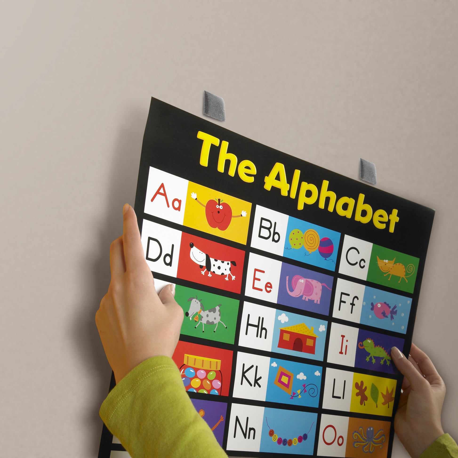 Ein Kind hält eine bunte Buchstabentafel mit passenden Bildern, die mit Scotch-FIX™ Ablösbare Montagequadrate 108-ST64-P (12,7 mm x 12,7 mm, 64 Stück/Packung) von 3M Deutschland GmbH an einer schlichten hellen Wand befestigt wurden.