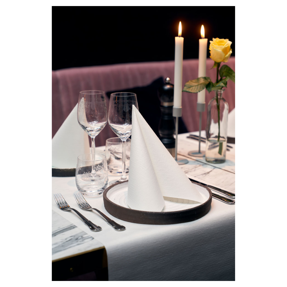 Ett matbord är elegant dukat med två Duni Dunilin®-servetter 48 x 48 cm Vit, vikta till trianglar på tallrikarna. Två stora brinnande ljus och en gul ros i en glasvas pryder bakgrunden. Vinglas, ett vattenglas och bestick kompletterar denna sofistikerade atmosfär med tyg-liknande charm.
