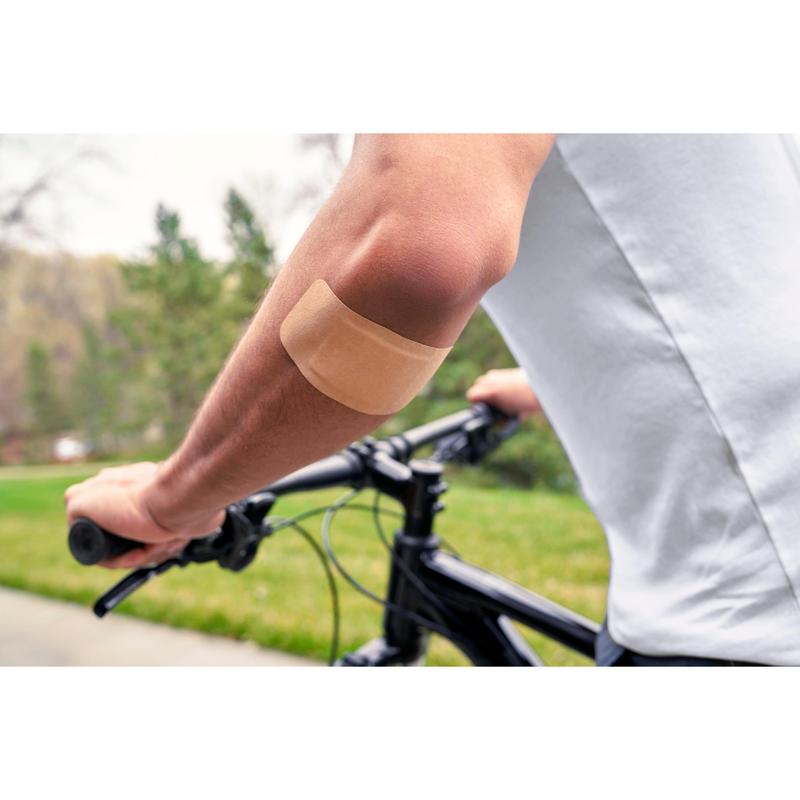 En person som bär en vit skjorta och cyklar utomhus har ett Nexcare™ Ultra Stretch MAXI Comfort Flexibelt Plåster (50 mm x 101 mm) från 3M Deutschland GmbH på den bara överarmen; i bakgrunden syns grönt gräs och träd.
