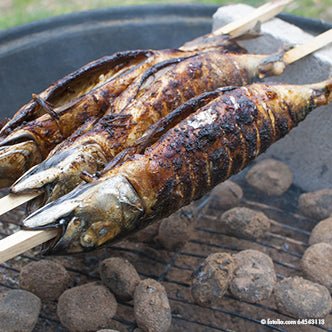 Två hela steckerlfiskar är uppträdda på PAPSTAR grillspett "pure" 80 cm (20 stycken) från PAPSTAR GmbH och grillas över glödande kol, där skinnet förkolnas och blir krispigt över den glödande kolen.