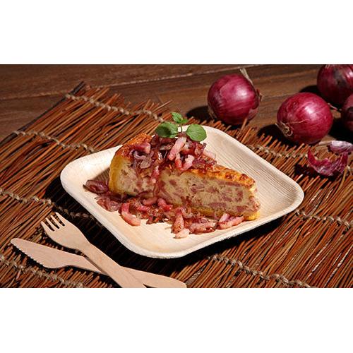 En skiva spansk tortilla med lök och örter serveras på en kvadratisk tallrik PAPSTAR 25, Palmblad "pure" från PAPSTAR GmbH, som vilar på en flätad matta med träbestick och rödlök i bakgrunden.