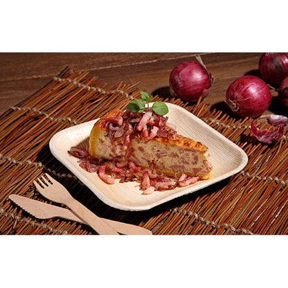 En skiva spansk tortilla med lök och örter serveras på en kvadratisk tallrik PAPSTAR 25, Palmblad "pure" från PAPSTAR GmbH, som vilar på en flätad matta med träbestick och rödlök i bakgrunden.