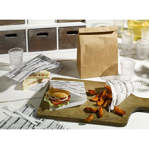 En smörgås, en hamburgare och potatisklyftor serveras i PAPSTAR 1000 spetsiga påsar (pergamentersättning, fettbeständig) från PAPSTAR GmbH, placerade på en träbräda med en brun papperspåse, genomskinliga koppar och plastbestick på ett vitt bord.