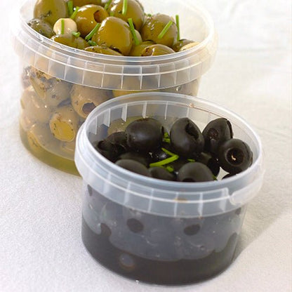 I två transparenta Starpak 30 delikatessbägare (770 ml, PP, Ø 11,8 cm · 11,3 cm) med originalförslutning finns oliver på vit bakgrund - i den bakre bägaren gröna oliver med örter och framme svarta oliver med örttillbehör.