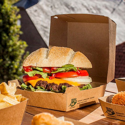 En cheeseburgare med sallad, tomat, lök och cheddarost ligger utomhus i en PAPSTAR GmbH Burgerbox i kartong (9x18,5x18,5 cm, ”100 % Fair”, 25 stycken). Pommes frites och andra tillbehör serveras separat bredvid.