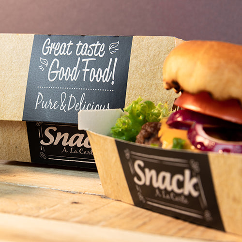 En kartongbricka med texten "Snack A La Carte" innehåller en hamburgare med sallad, tomat och lök. I bakgrunden står en PAPSTAR 50 burgerlådor, kartong "pure" "Good Food" från PAPSTAR GmbH: God smak Good Food! Pure & Delicious.
