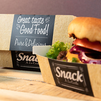 En kartongbricka med texten "Snack A La Carte" innehåller en hamburgare med sallad, tomat och lök. I bakgrunden står en PAPSTAR 50 burgerlådor, kartong "pure" "Good Food" från PAPSTAR GmbH: God smak Good Food! Pure & Delicious.