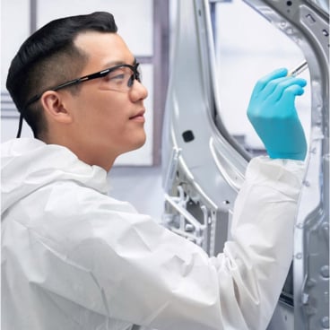 Eine Person mit Schutzbrille, blauen Handschuhen und einem KleenGuard® KGA20 Atmungsaktiver Schutzanzug mit Kapuze von Kimberly-Clark GmbH benutzt ein Werkzeug an einer Metallstruktur in einer Industrie- oder Laborumgebung.